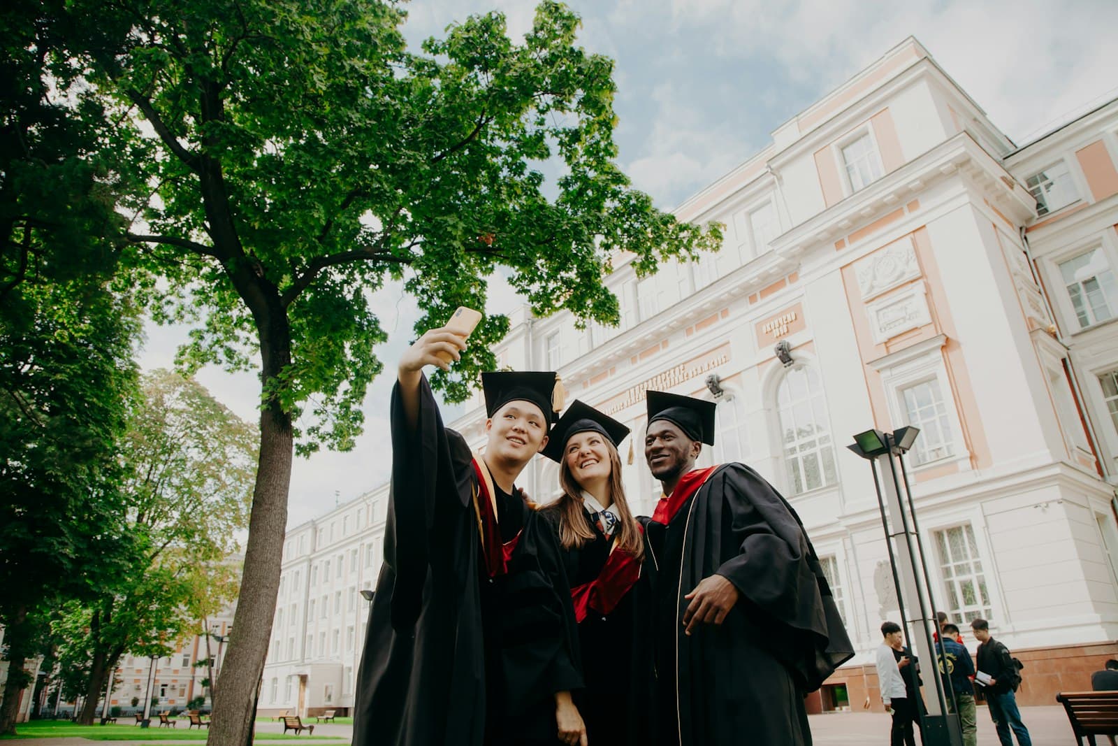 Three graduates in caps and gowns taking a selfie in front of their university