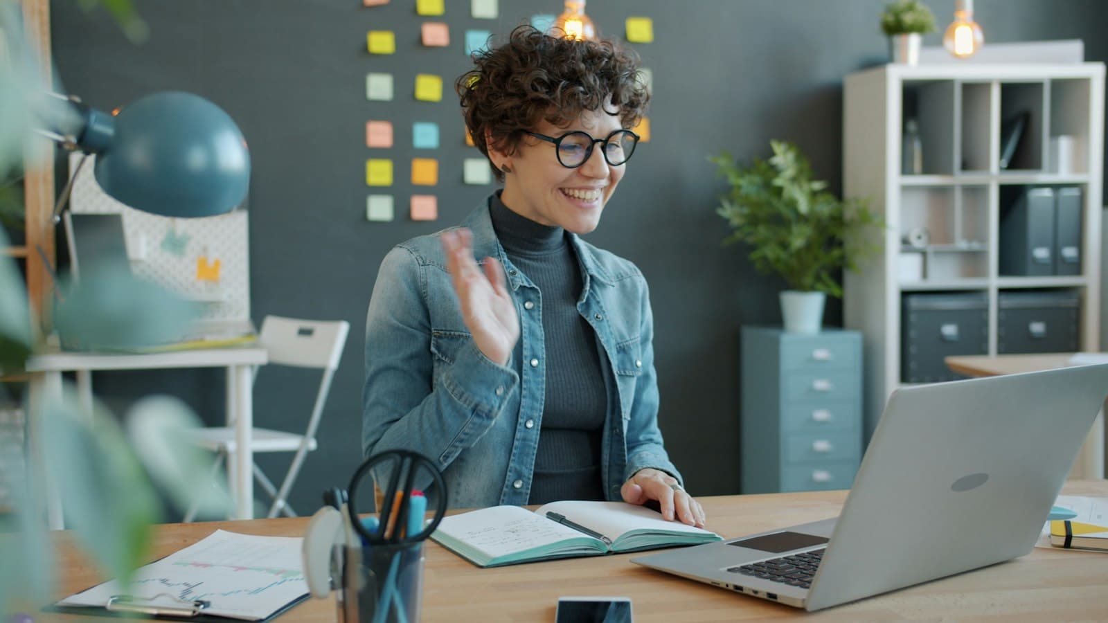 A woman waving hello on a video call at her desk